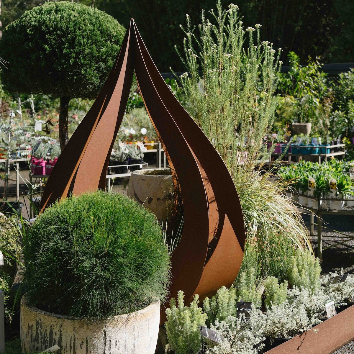 A rusted steel garden sculpture with three upward-curving angular blades, displayed in a lush garden setting with potted greenery and foliage in the background.
