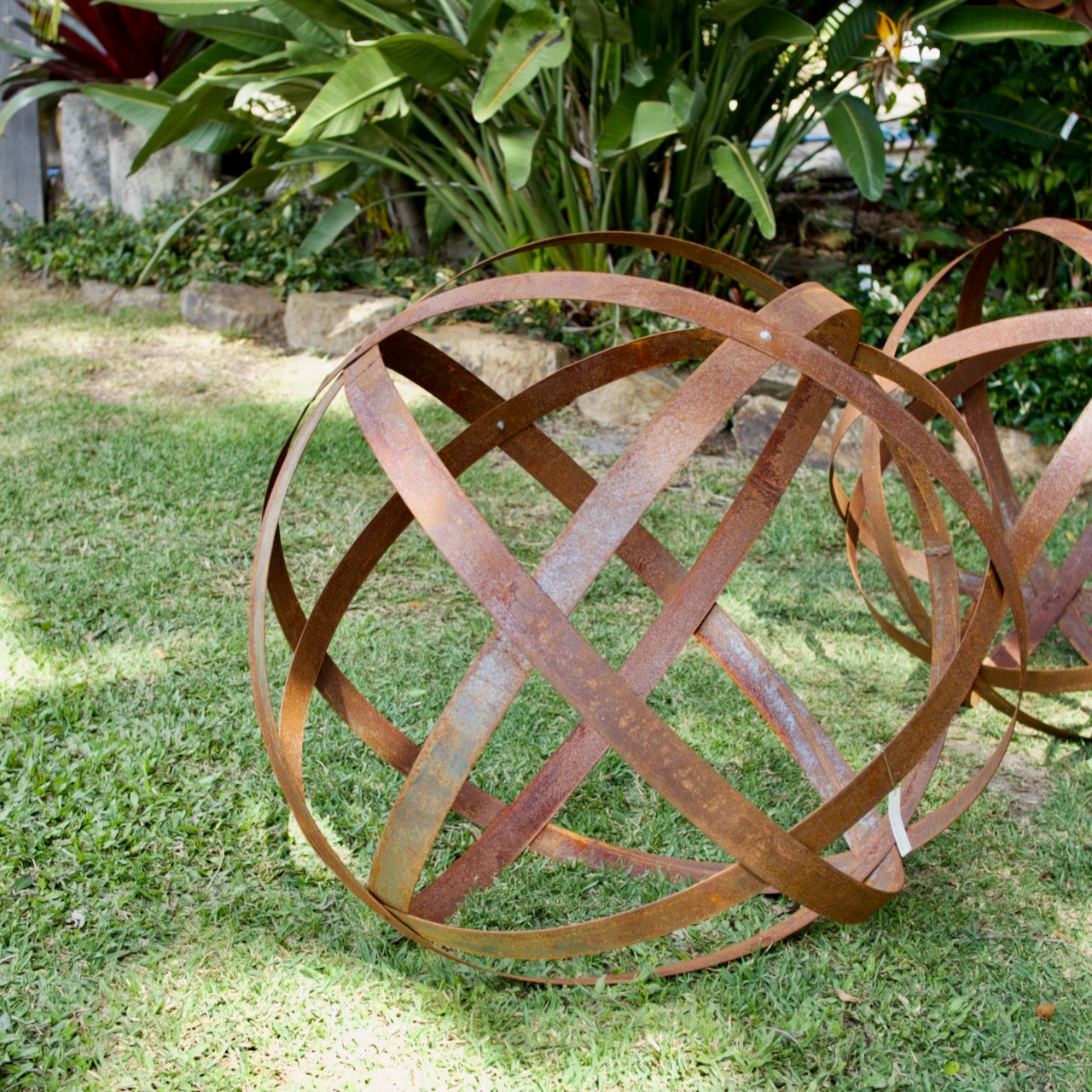 A rusted metal decorative sculpture in the shape of interlocking rings or a lattice sphere, placed on a green grass lawn with tropical plants in the background.
