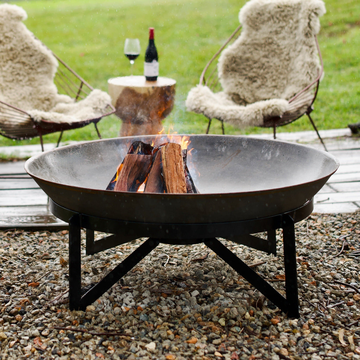 Fire pit with flames in a garden setting with two cushioned chairs and wine glasses.