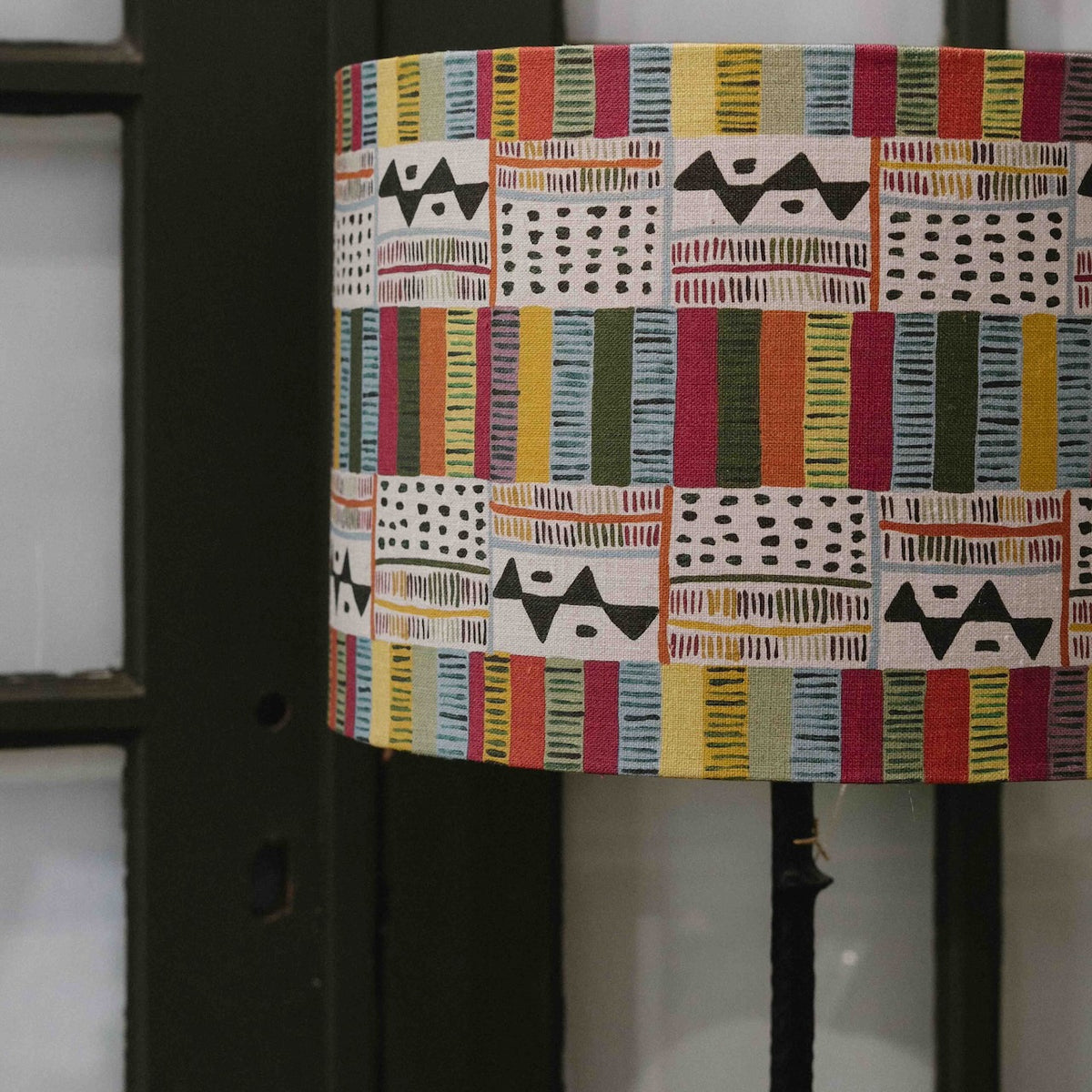 A close-up of a cylindrical lampshade featuring a colorful patchwork pattern with geometric and striped textile designs in red, yellow, blue, green, black, and white, shown against a dark background.