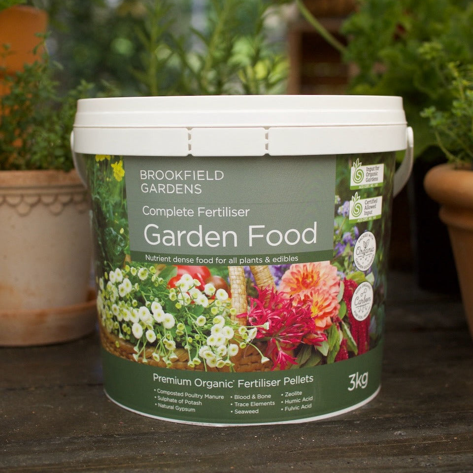 A 3kg tub of Brookfield Gardens Complete Gardeniser Food placed on a dark wooden surface with a blurred background of potted green plants.