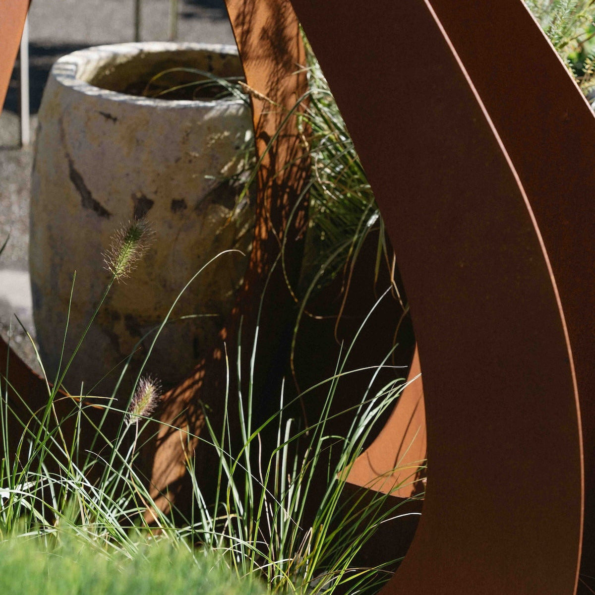 Corten steel yard sculpture in a garden setting, featuring rusted brown curved metal panels among green grass and a stone planter.