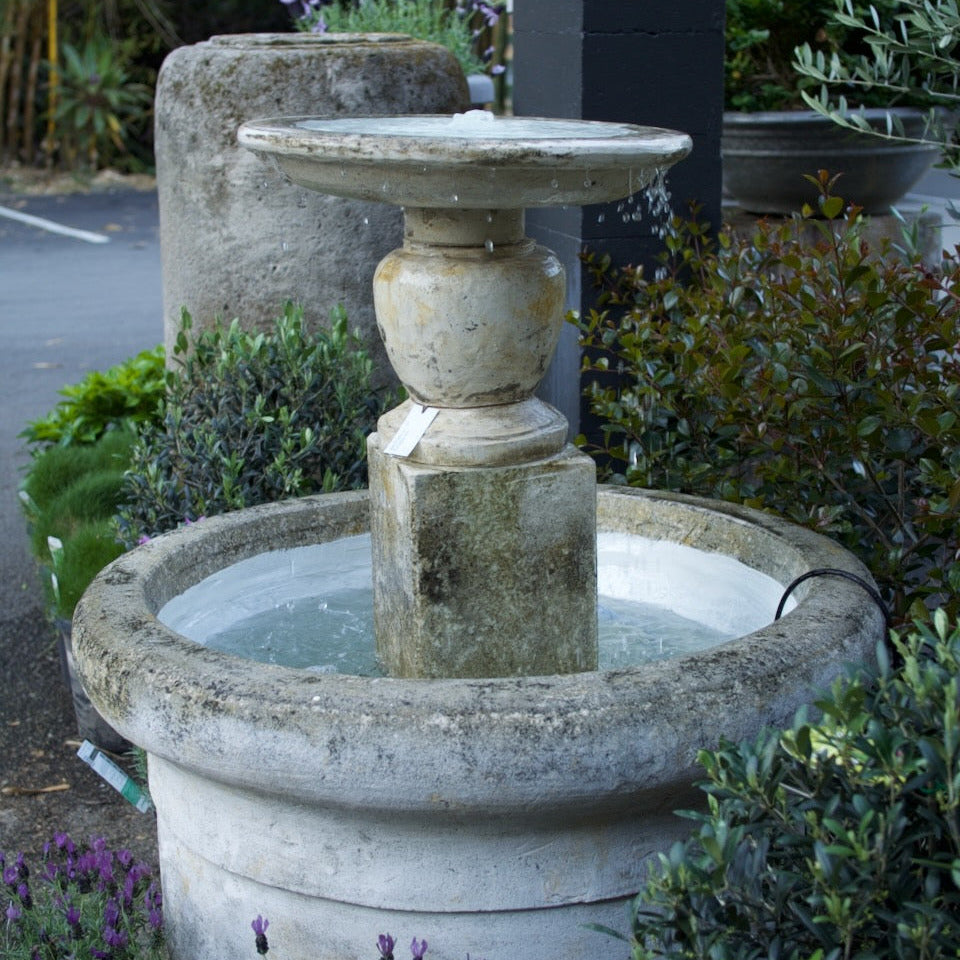 A round stone fountain with a central pedestal and shallow basin, placed outdoors among green shrubs and small purple flowers.