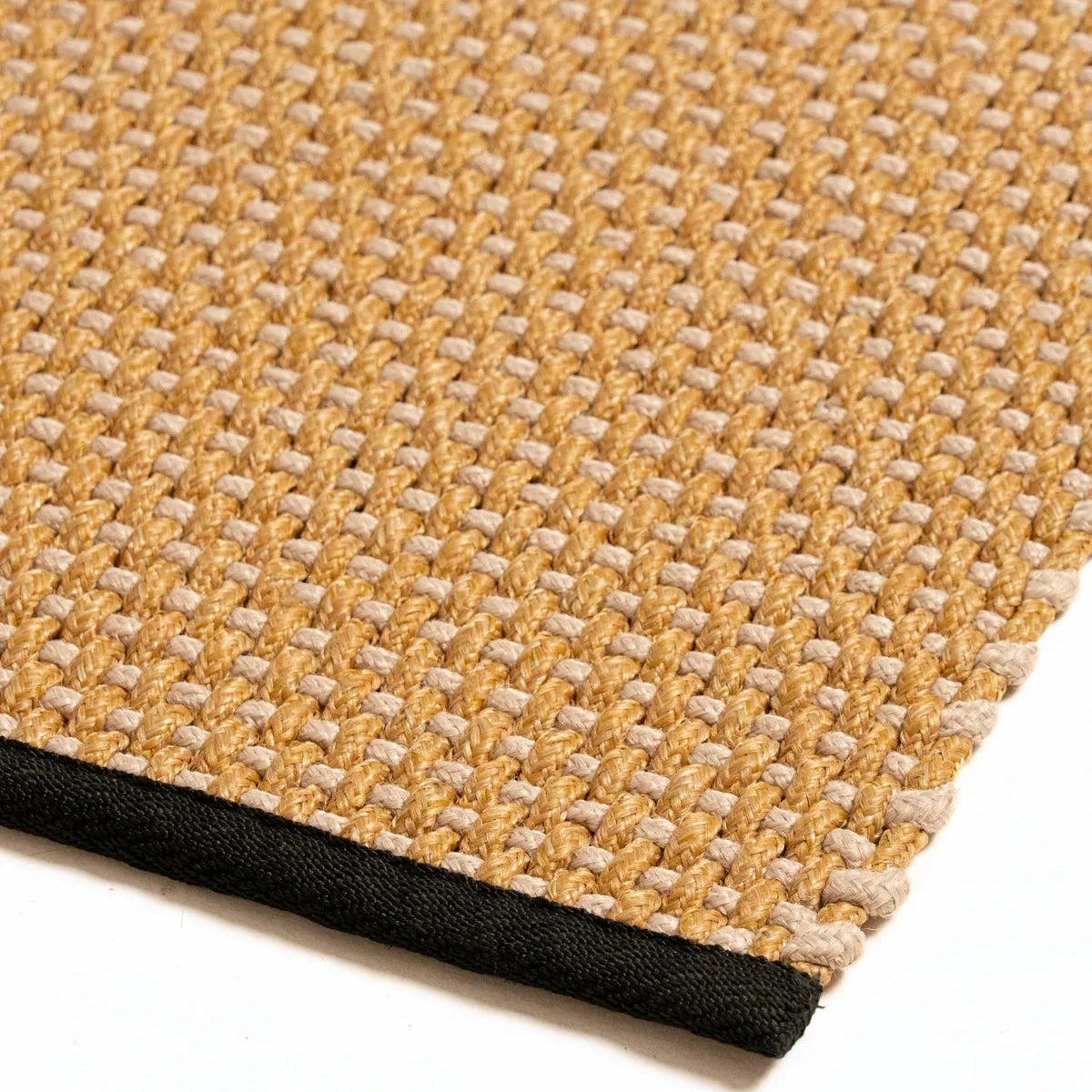 Close-up of a woven rug corner showing a tan and cream textured pattern with a black fabric edge on a white background.