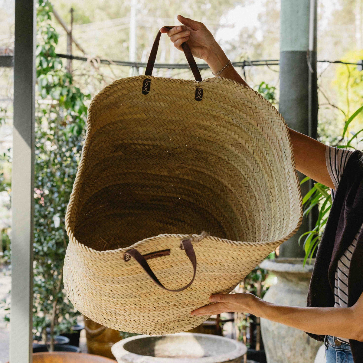A large woven straw tote bag with dark brown leather handles, held by a person in a rustic, sunlit outdoor setting with green plants in the background.