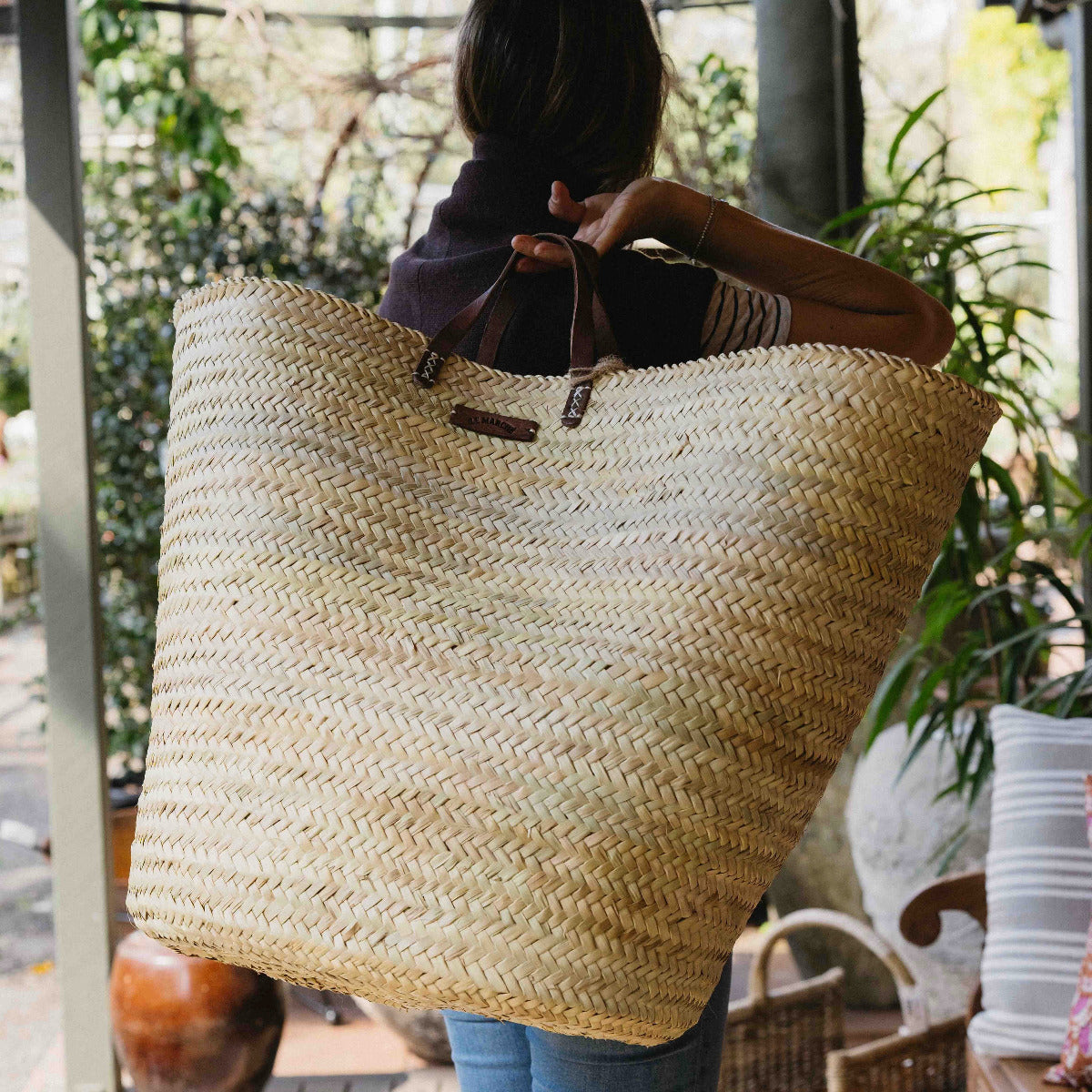 A person carrying a large natural straw tote bag with dark brown leather handles in an outdoor patio setting.
