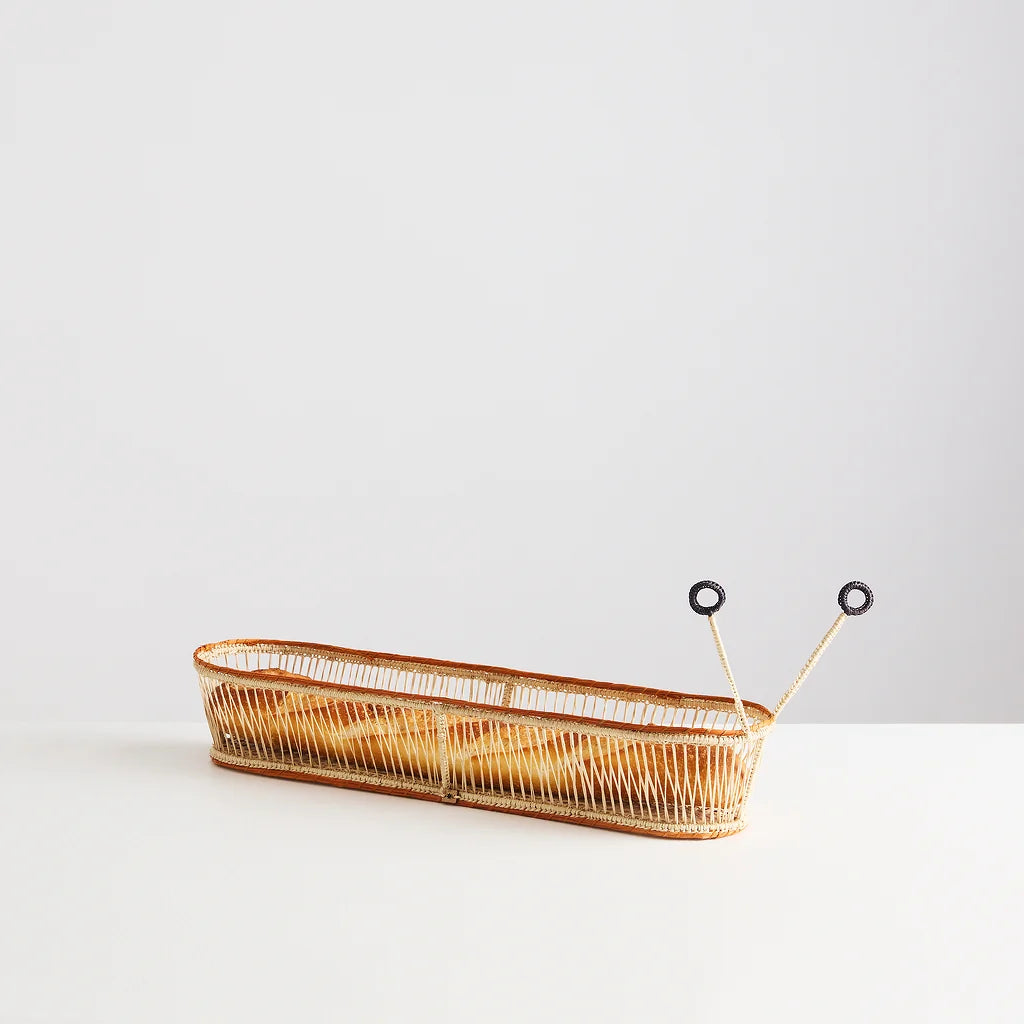 A long, narrow woven basket with a natural tan and brown finish, featuring two small black-and-white snail eyes on thin sticks extending from one end, photographed on a clean white surface against a light gray background.