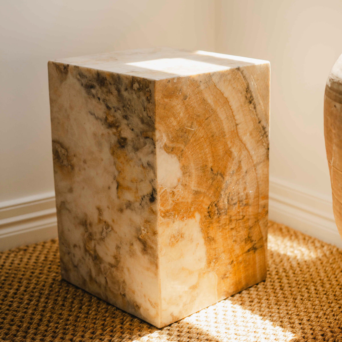 Natural onyx stone pedestal cube with honey and cream veining placed on woven floor mat indoors.