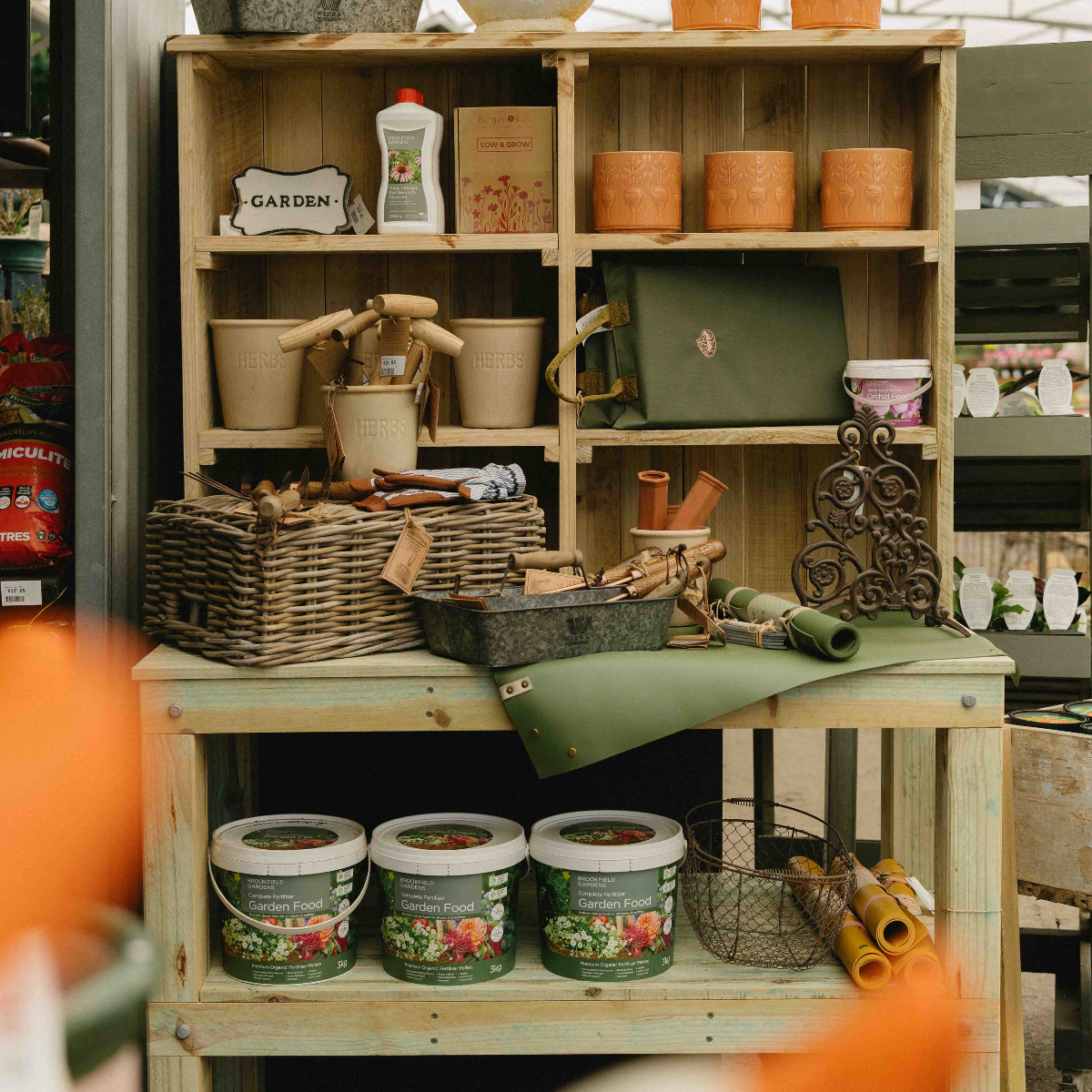A rustic wooden display shelf filled with gardening and home decor items, including terracotta pots, wicker baskets, a green tote bag, metal tools, and three labeled garden soil buckets in the foreground.