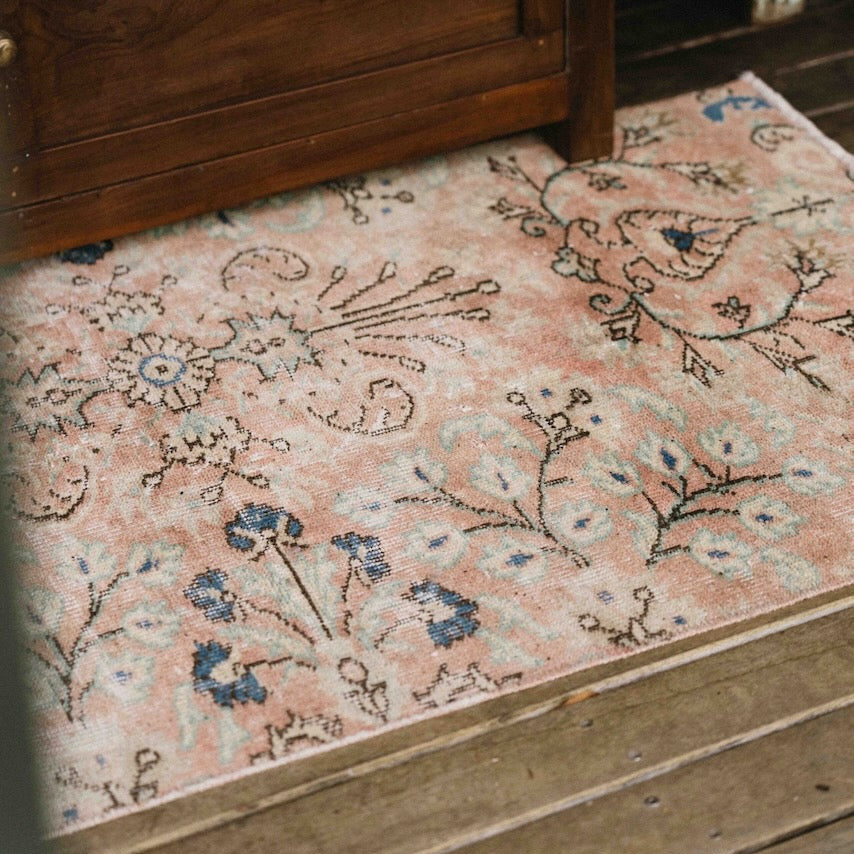 A close-up of a vintage-style area rug with a dusty pink background and blue floral motifs, shown on wooden steps next to a dark wood cabinet.