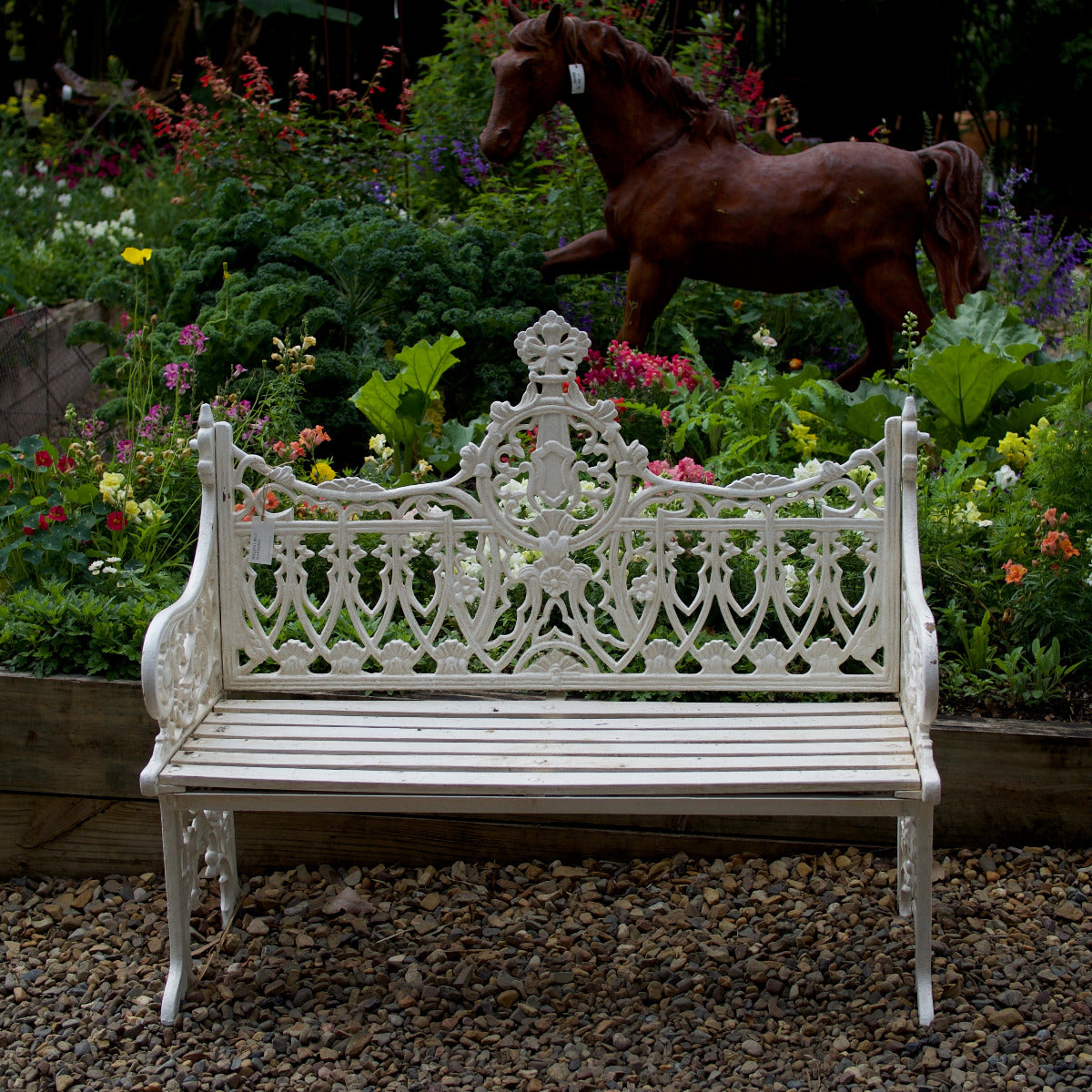 White ornate Shelly garden bench with decorative detailing displayed amongst lush flowering plants at Brookfield Gardens Brisbane.