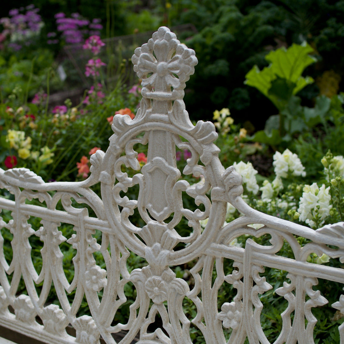 White ornate cast iron garden bench with intricate scrollwork and a decorative crest finial, set against a lush green garden background with colorful flowers.