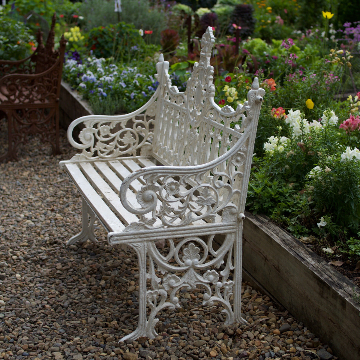 A white ornate cast iron garden bench with decorative scrollwork and a slatted backrest, placed on a gravel path in a flower garden.