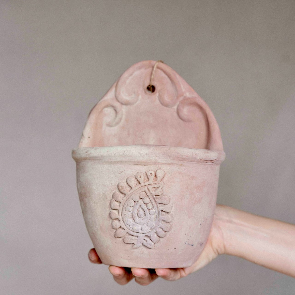 A hand holding a dusty pink terracotta hanging planter with a decorative embossed paisley design on the front and a hanging loop at the top, set against a plain gray background.