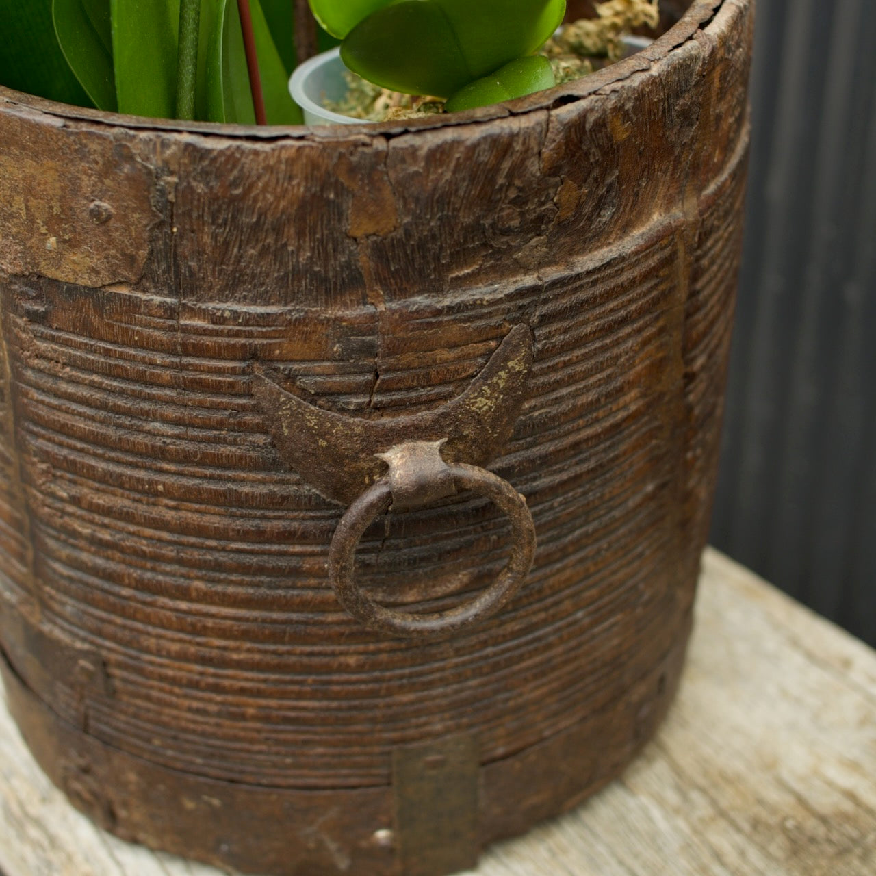 A rustic, cylindrical wooden planter with horizontal grooves and a weathered metal ring handle, shown holding green plants on a light wooden surface.