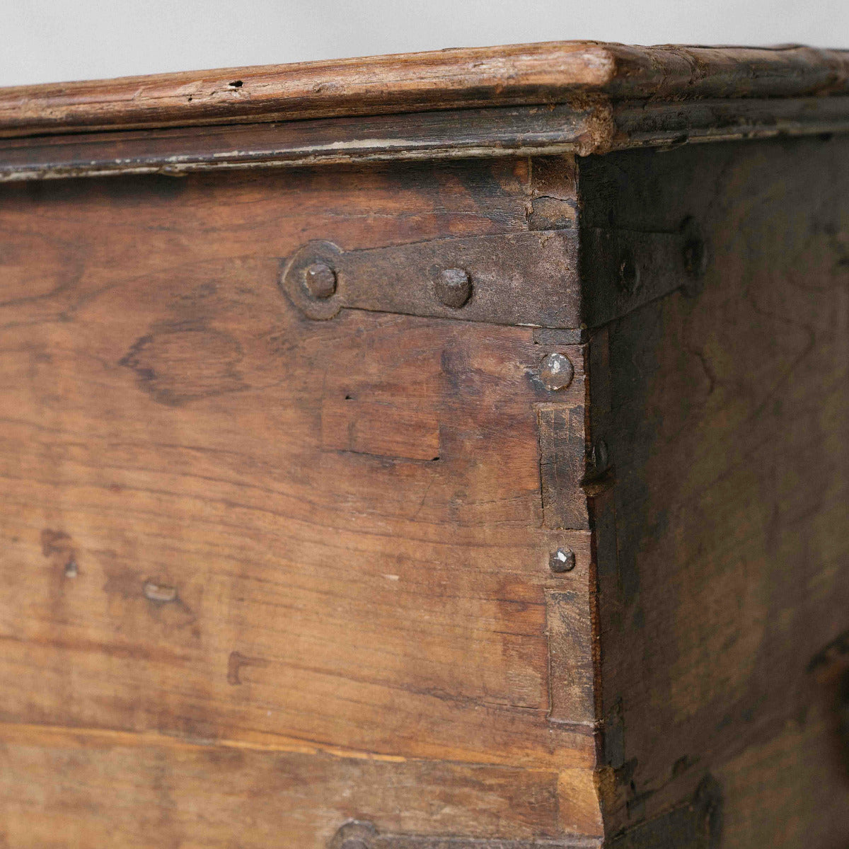 Close-up of the corner of a rustic wooden trunk or chest with metal corner brackets and rivets, showing weathered wood grain and aged finish.
