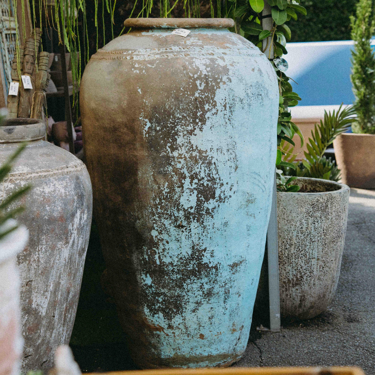 A large distressed ceramic vase with a weathered turquoise and brown patina, displayed outdoors among other potted plants and garden decor.