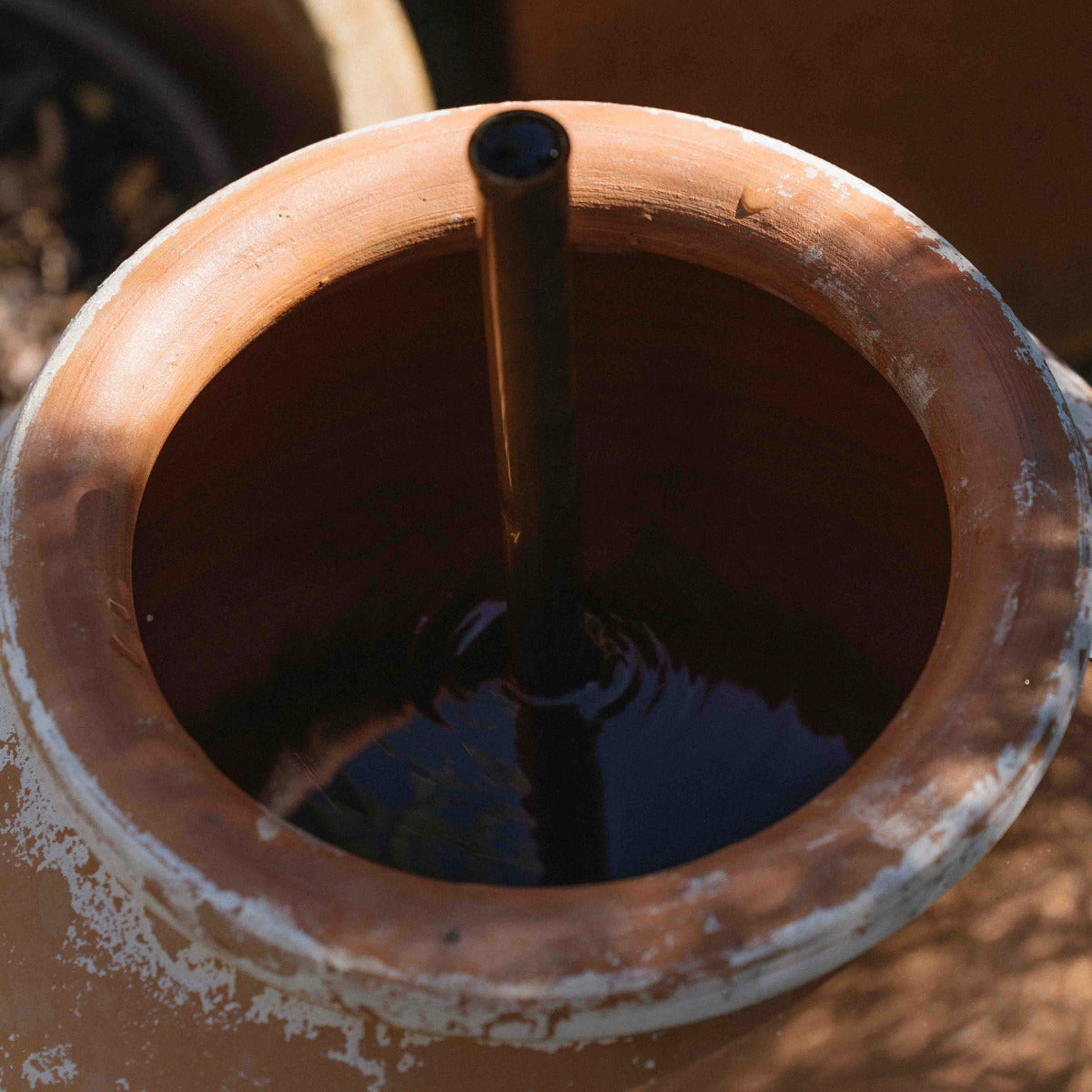 A close-up of a terracotta clay water container with a dark metal tube inserted into the water surface.
