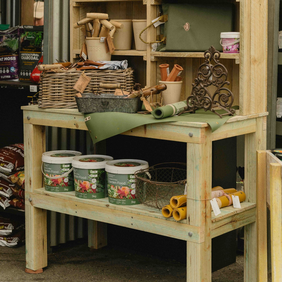 A light natural wood garden workbench with shelves, styled with gardening tools, pots, and rolled green fabric, set in a garden center or workshop environment.
