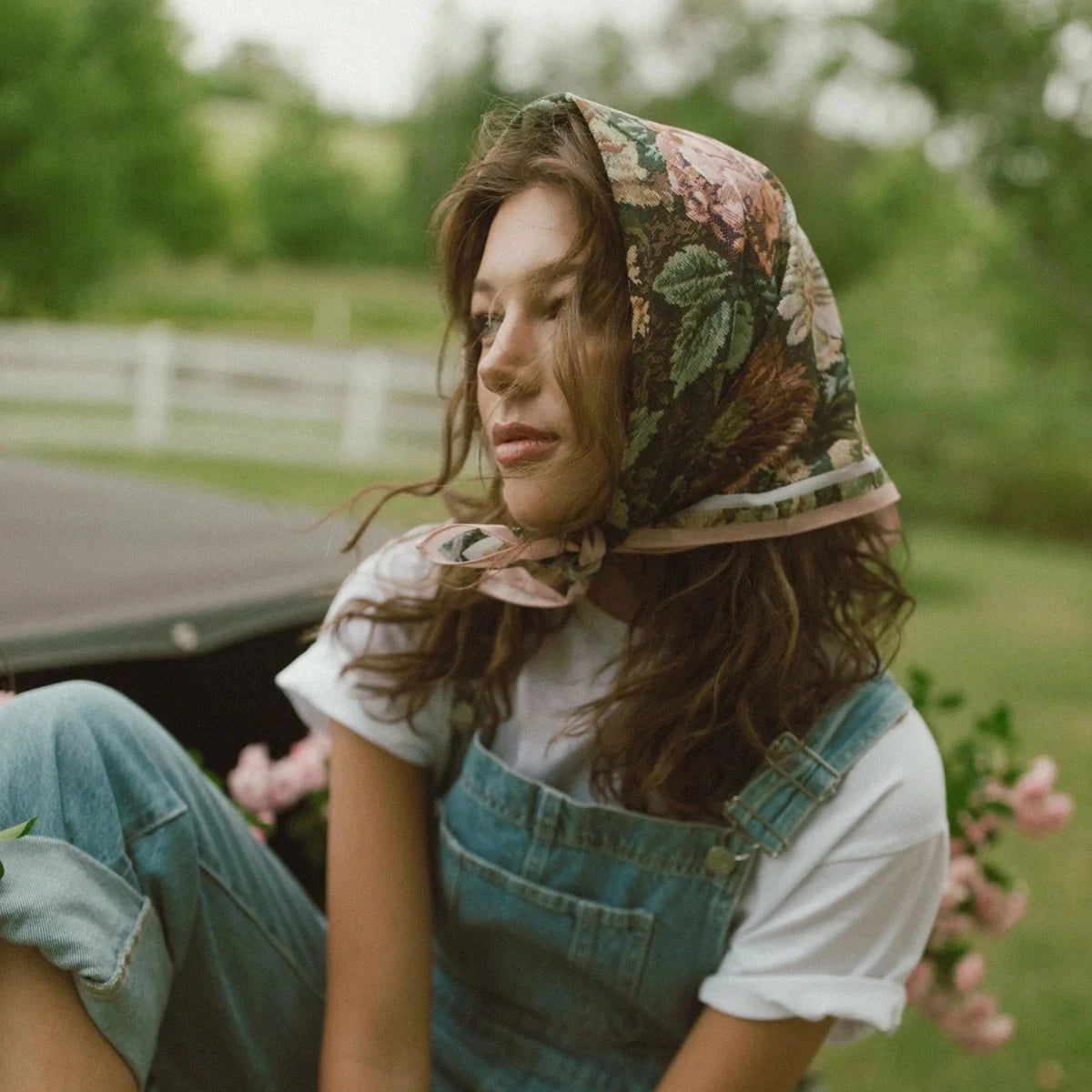 Woman wearing a floral headscarf and denim overalls sitting outdoors.