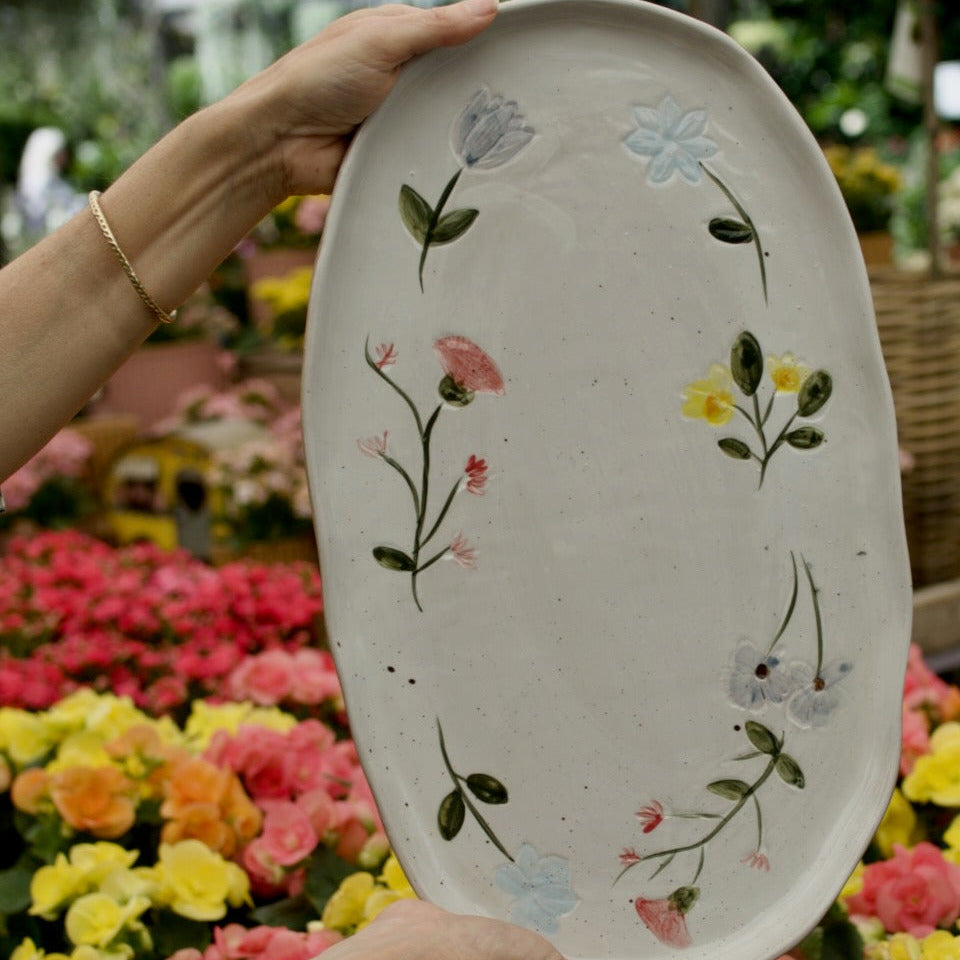A person holds an oval white ceramic serving platter decorated with hand-painted floral designs in green, pink, yellow, and light blue, set against a blurred background of a vibrant flower garden.
