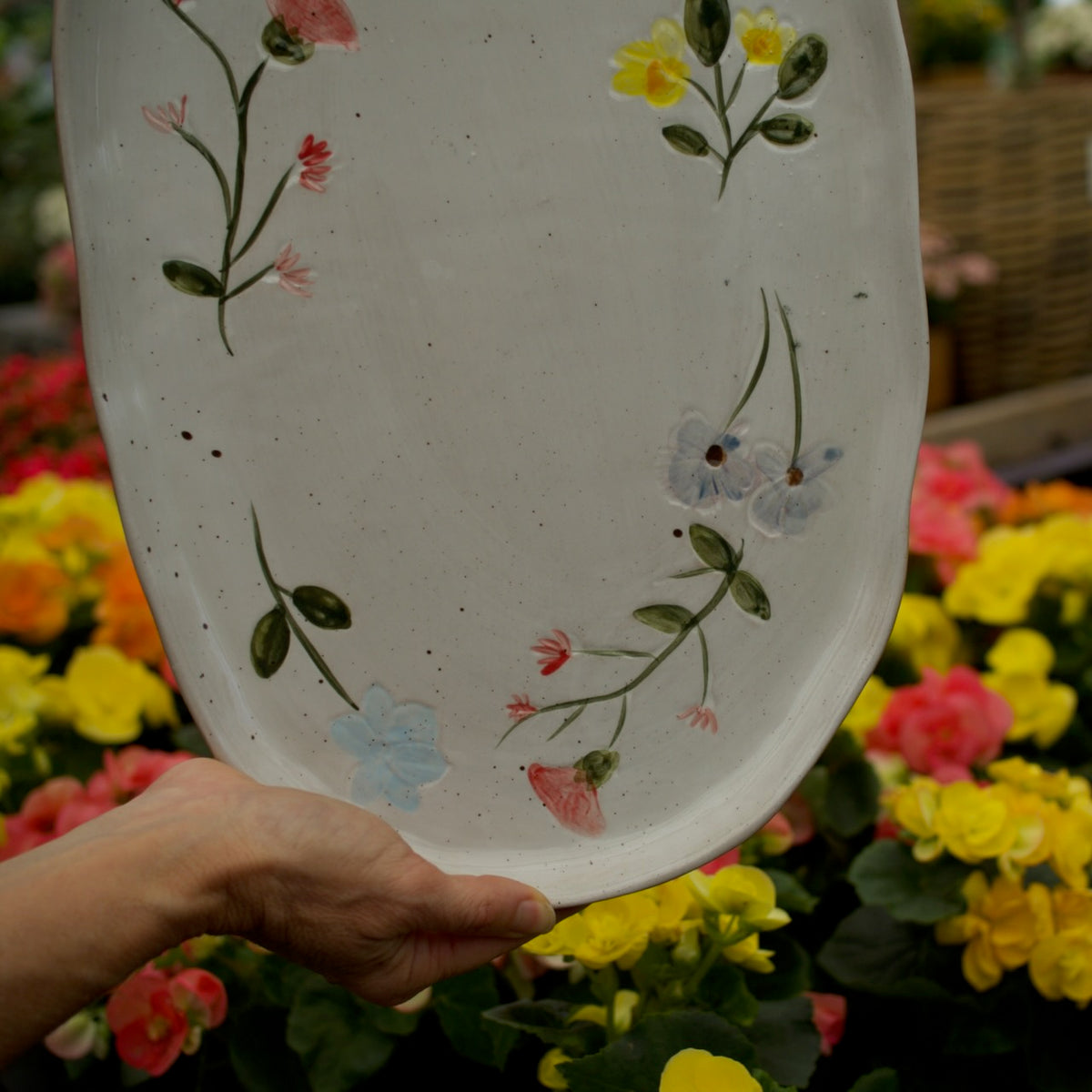 A hand holds a large, oval-shaped white ceramic plate or planter decorated with hand-painted green vines and small flowers in yellow, pink, and light blue, set against a background of colorful flowers.
