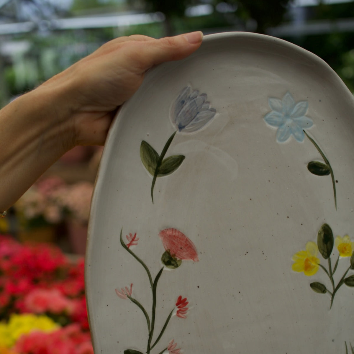 A hand holds a large oval ceramic serving platter with a speckled off-white glaze and hand-painted floral designs, set against a blurred greenhouse background with colorful flowers.