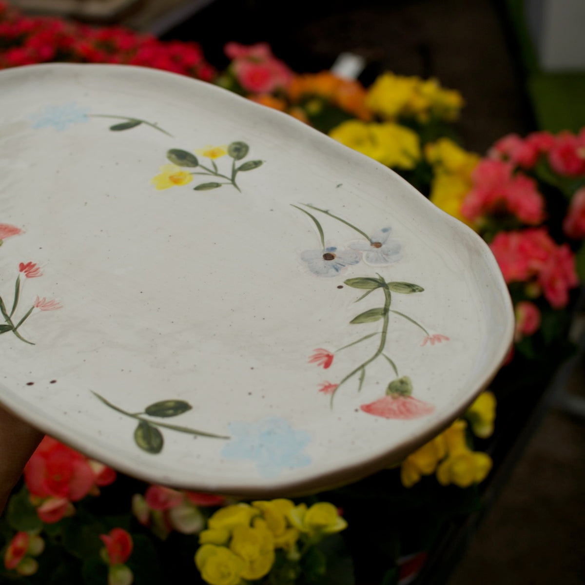 A close-up of a hand-painted ceramic serving platter with a white glaze and delicate floral designs, held in front of a blurred background of colorful flowers.
