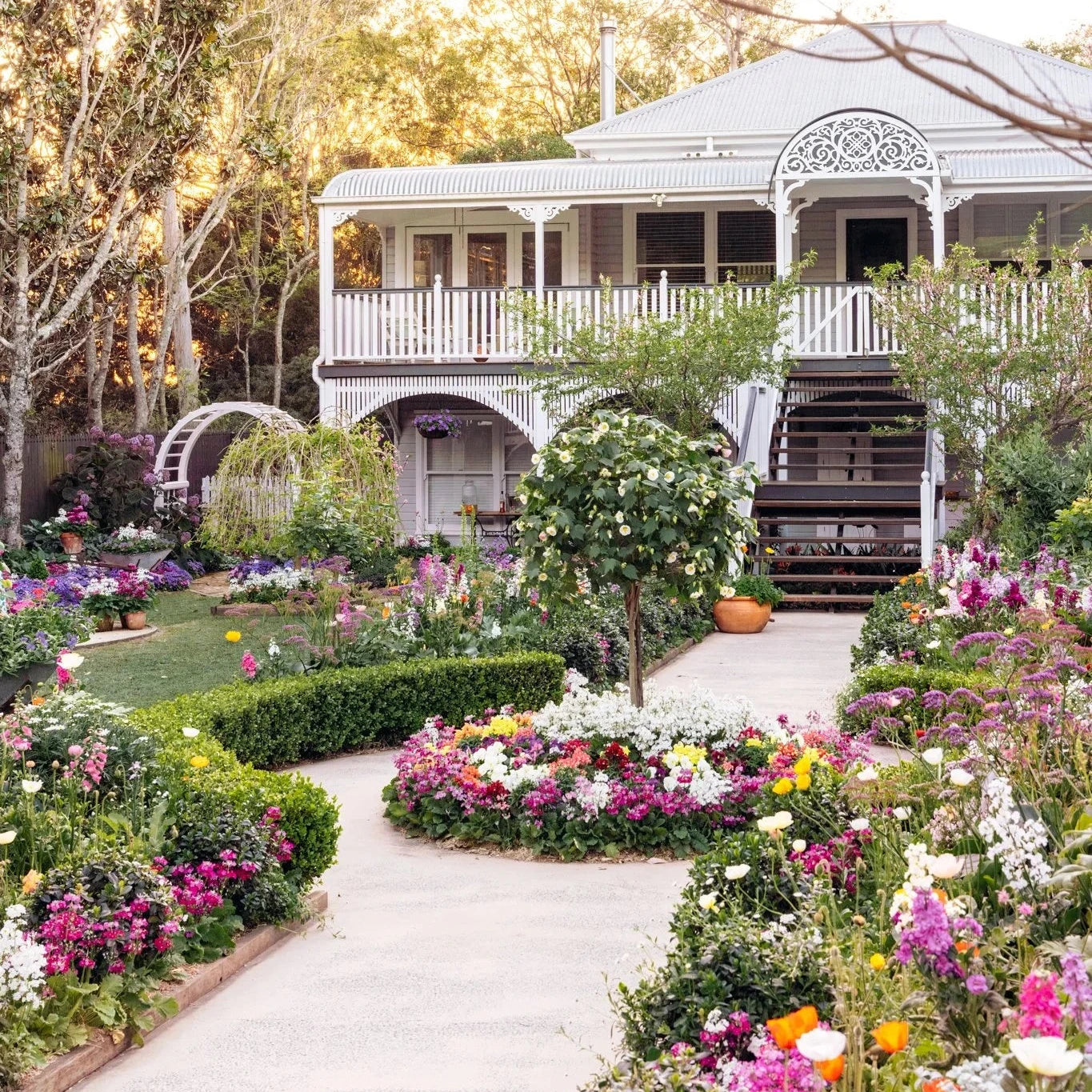 A beautifully landscaped garden pathway leading up to a large white Victorian-style house with a wraparound porch, surrounded by lush greenery and colorful flower beds.