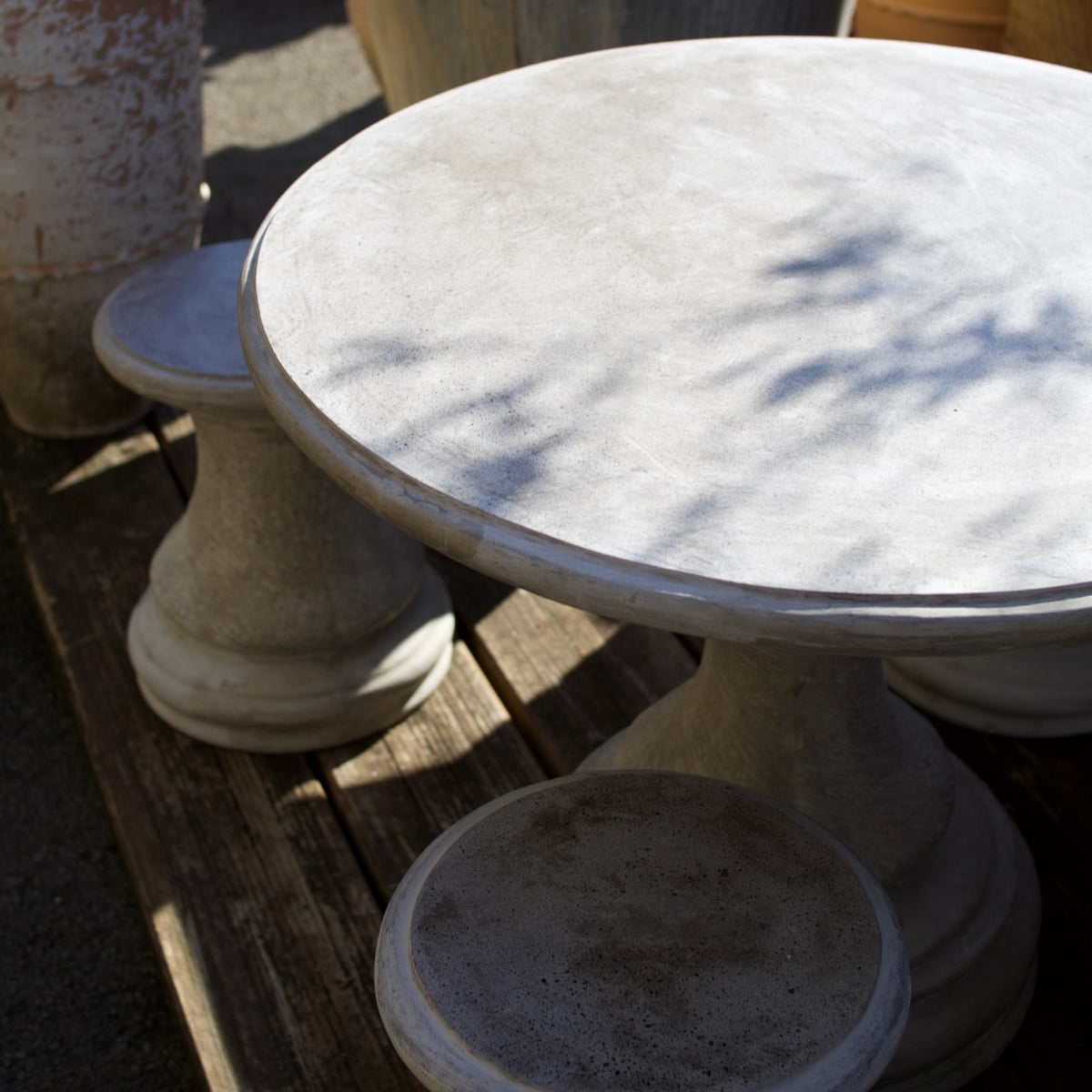A round concrete pedestal table with matching stone stools, photographed outdoors on a wooden deck with dappled sunlight and shadows across the tabletop.