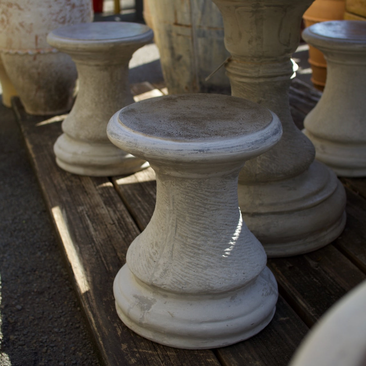 A distressed white stone garden pedestal stool with a rustic weathered finish, shown on a wooden deck alongside other similar stone planters and stools.