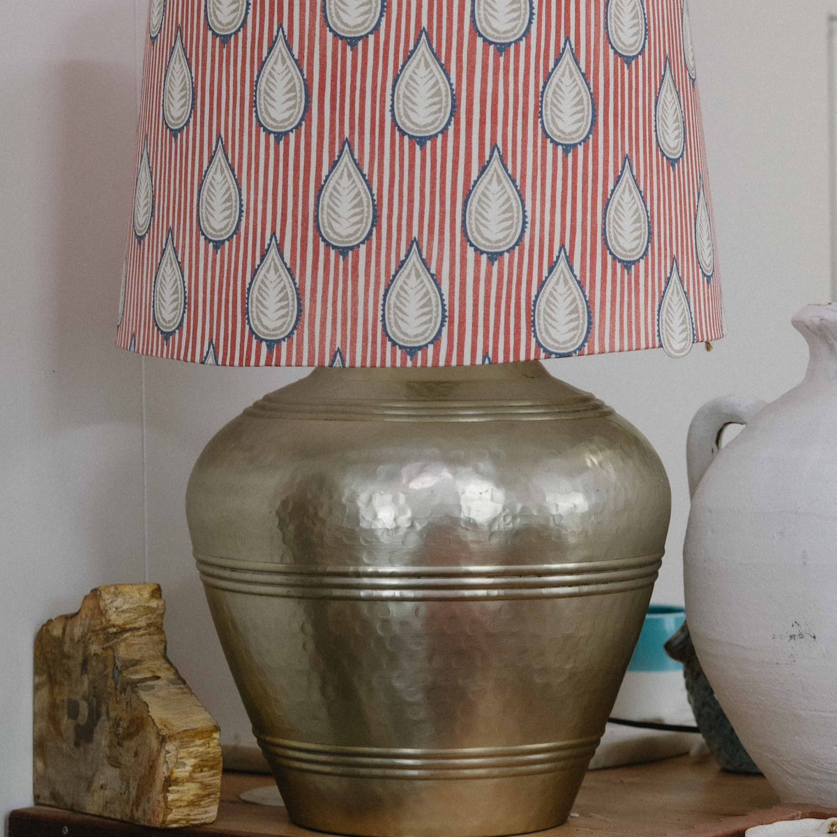 A table lamp with a hammered metallic gold base and a patterned drum shade featuring vertical red stripes and white teardrop motifs outlined in blue, shown on a shelf next to a white ceramic jug.
