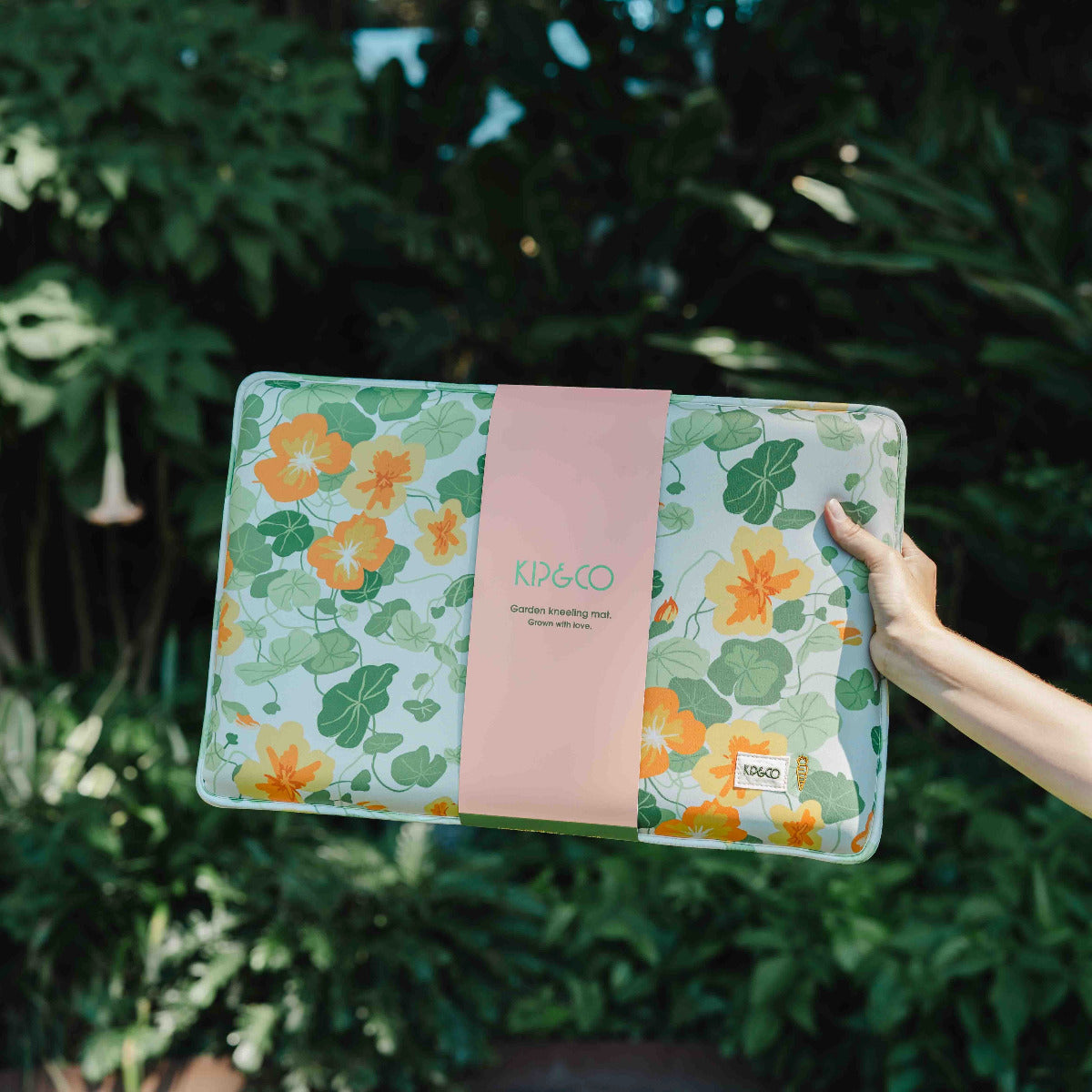 Floral-patterned product with 'Kip & Co' branding held by a hand against a green leafy background
