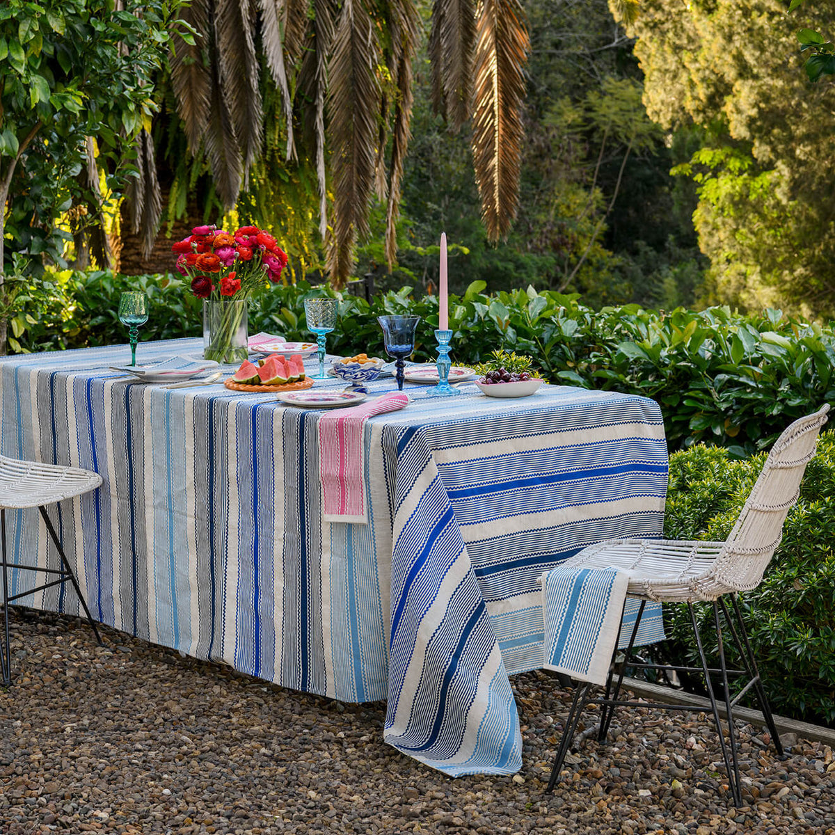 A blue and white striped tablecloth draped over an outdoor dining table set with glassware, candles, and a red flower arrangement, surrounded by chairs and lush garden foliage.