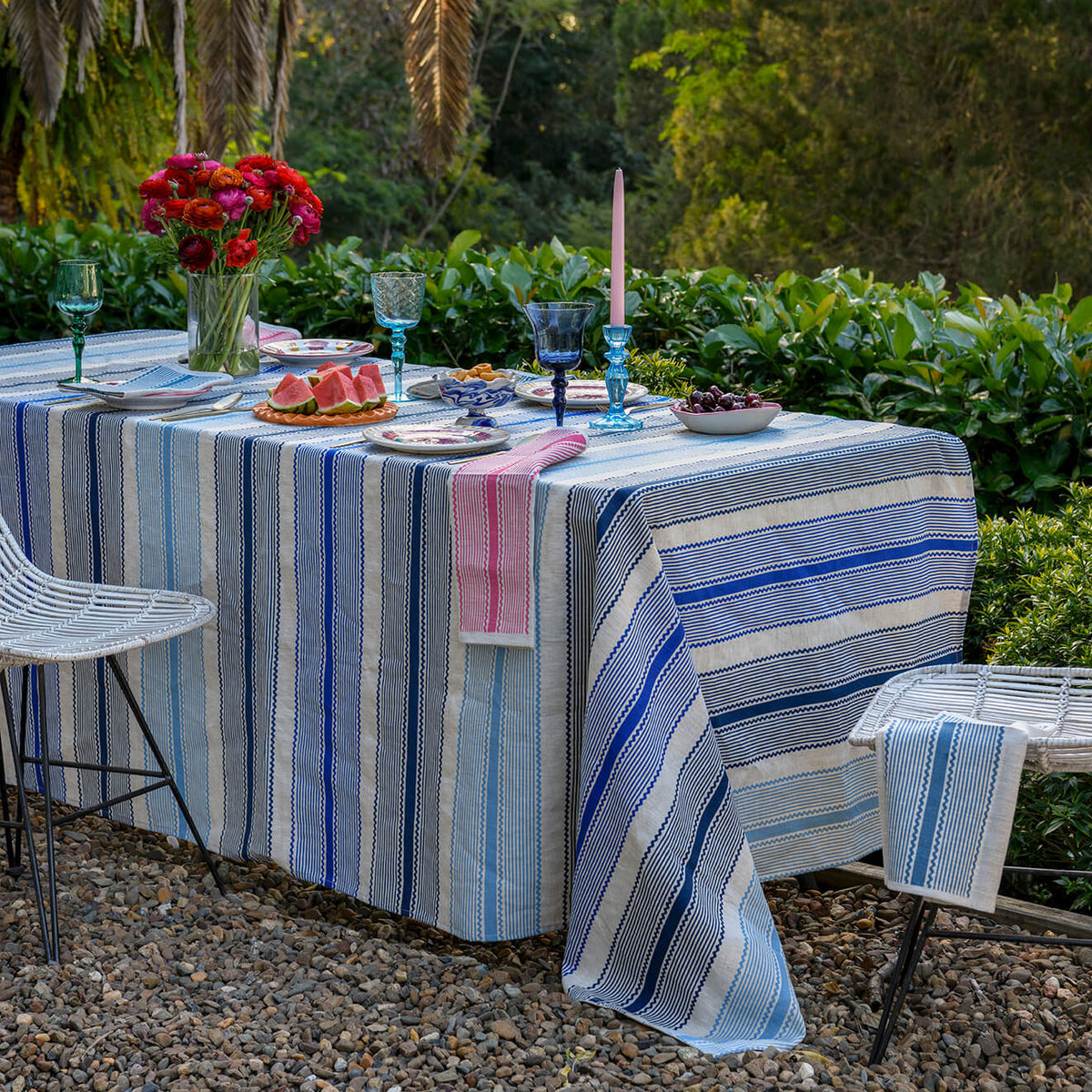 A blue and white striped tablecloth draped over an outdoor dining table set with plates, glasses, and a pink napkin, surrounded by chairs in a lush garden setting.