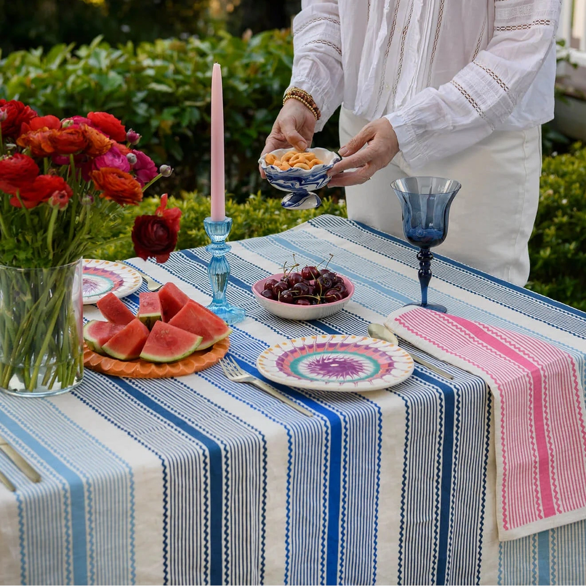A woman in a white blouse and wide-brim straw hat serves food from a small bowl at an outdoor dining table set with blue and white striped table linens, colorful plates, watermelon, flowers, and pink candles.