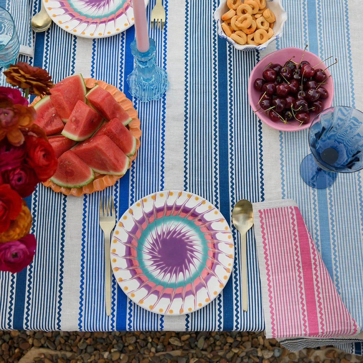 A styled outdoor table setting with a blue and white striped tablecloth, two matching patterned plates, bowls of watermelon, pretzels, cherries, pink napkins, and blue glassware.