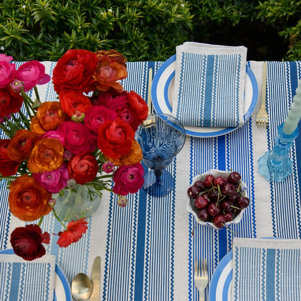 A styled outdoor table setting with blue and white striped tablecloths, matching napkins, blue glass goblets, silver cutlery, a large bouquet of red, pink, and orange roses, and a bowl of cherries.