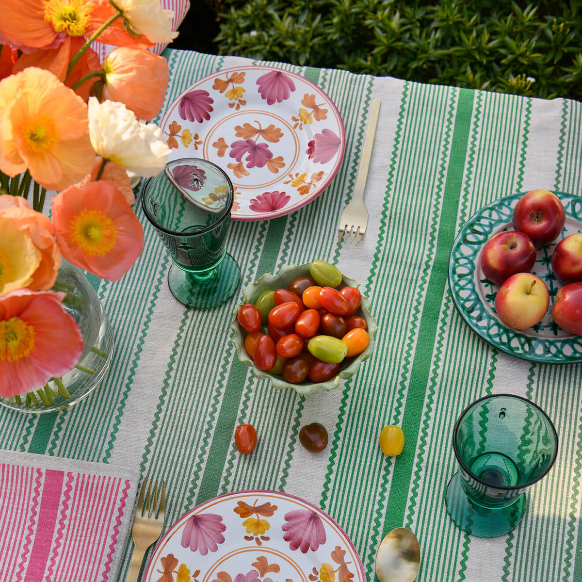 A styled outdoor table setting with a green and white striped tablecloth, floral plates, green glassware, a pink napkin, and a bowl of cherry tomatoes, with flowers in the upper left corner.