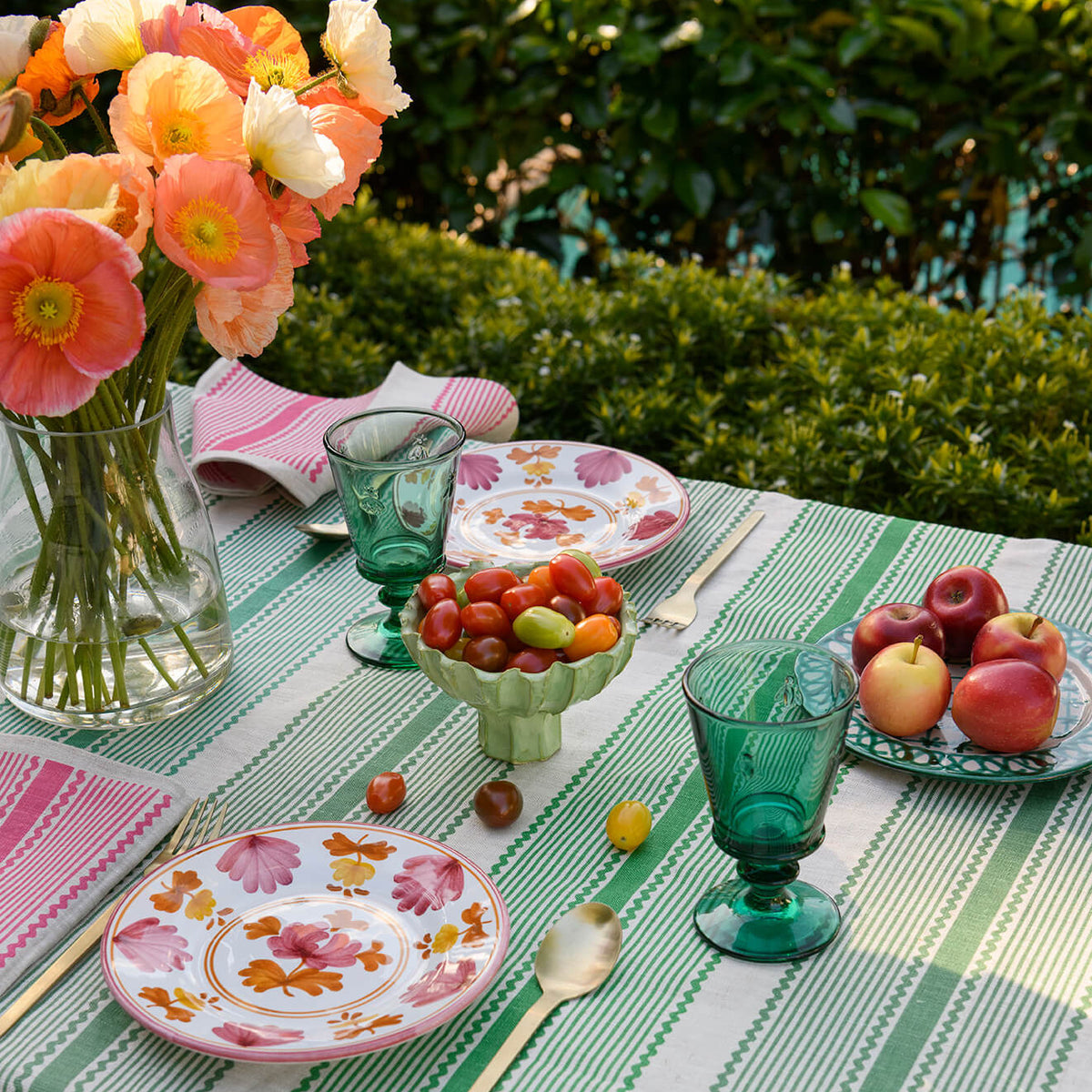 A styled outdoor dining table with a green and white striped tablecloth, floral plates, green glass goblets, pink napkins, a vase of orange flowers, and fruit on a garden table setting.