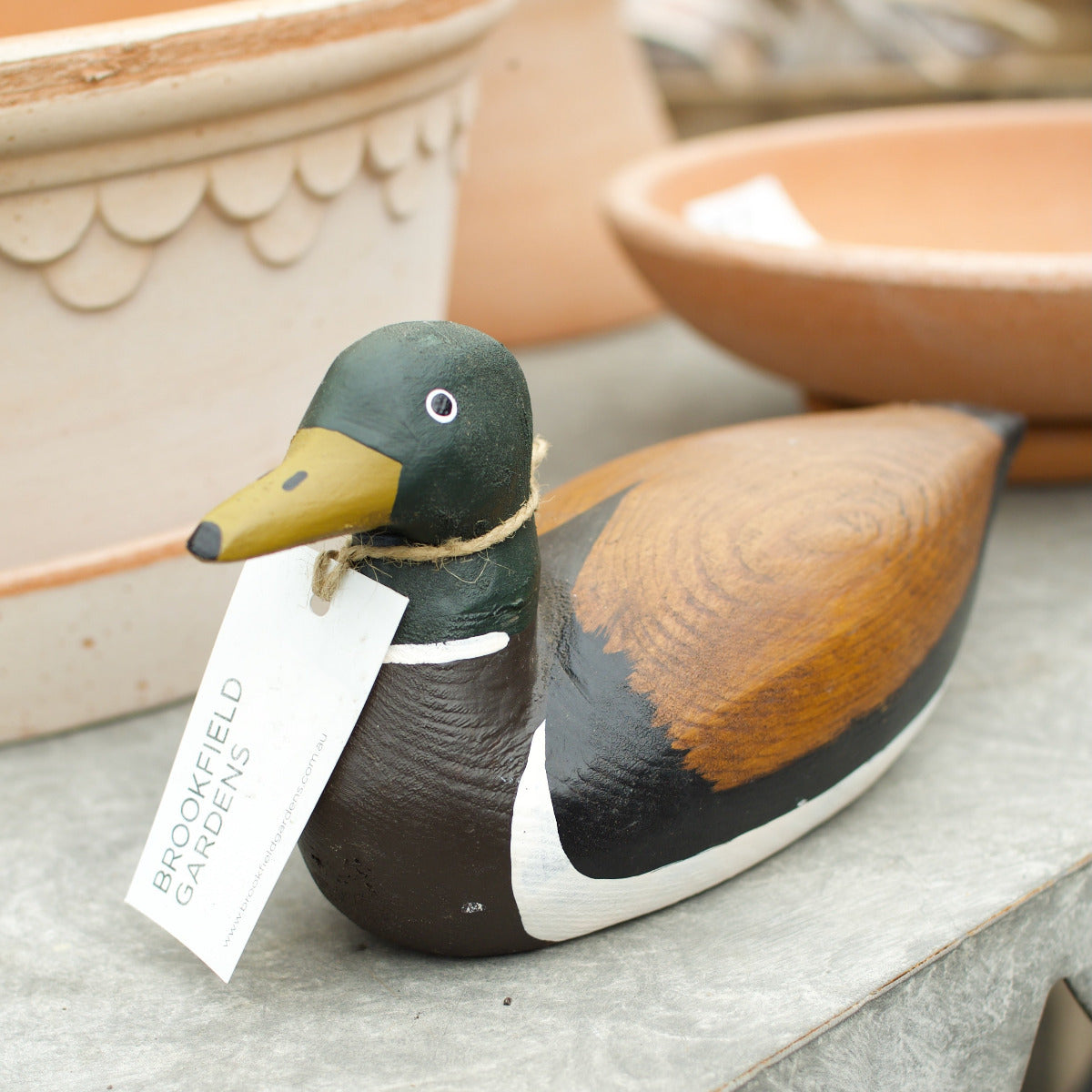 A hand-painted wooden mallard duck ornament with a white hanging tag reading 'BROOKFIELD GARDENS', set on a gray stone surface with terracotta pots in the background.