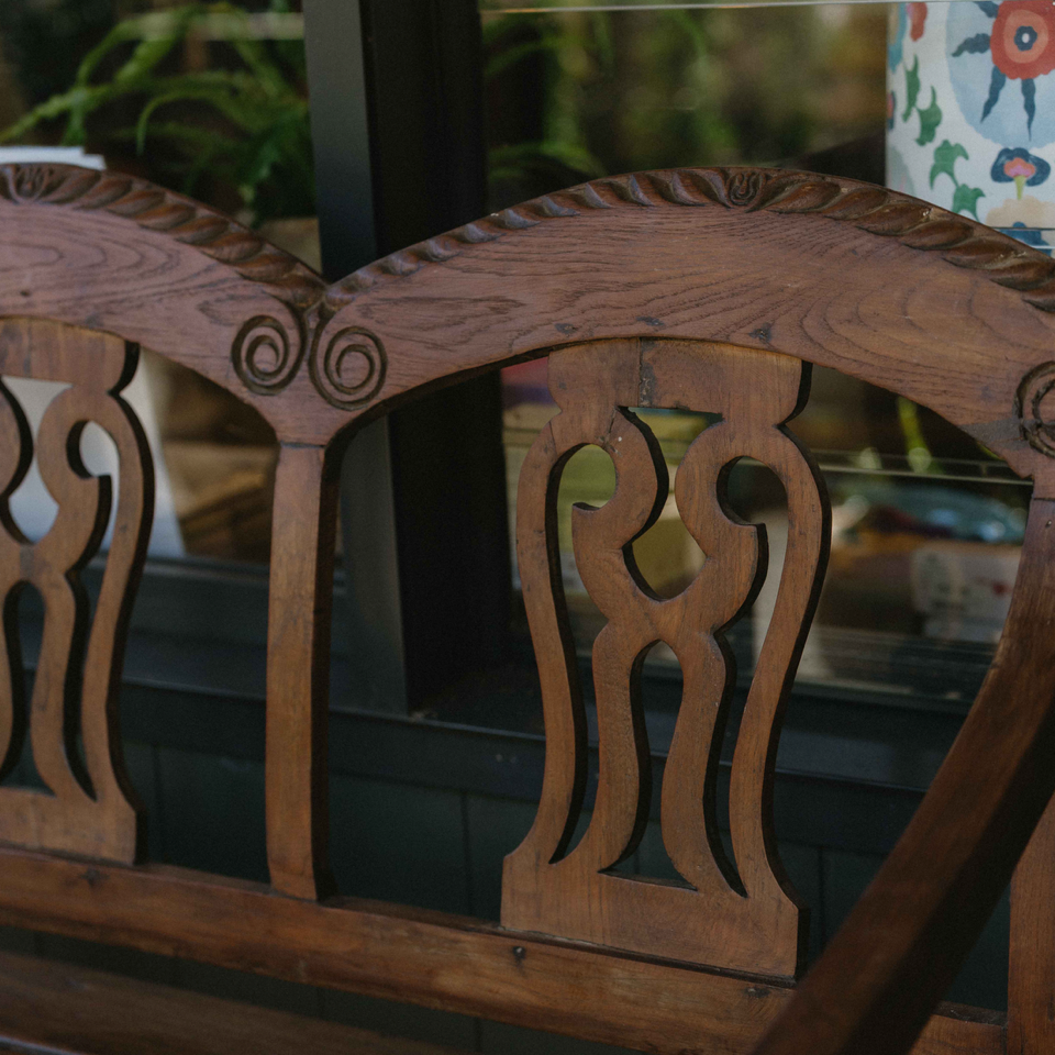Close-up of a carved wooden chair back with ornate scroll and curved cutout details, shown in warm medium brown wood tones.